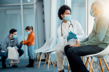 Black female doctor talking to senior man in a waiting room at medical clinic during coronavirus pandemic. 