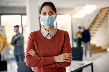 Confident businesswoman wearing protective face mask while working in the office. There are people in the background. 