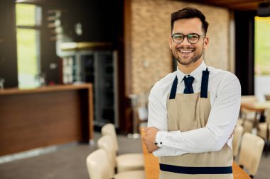Young happy waiter standing with arms crossed in a cafe and looking at camera.