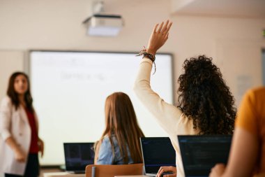 Rear view of high school student raising her arm to ask a question during lecture in the classroom.
