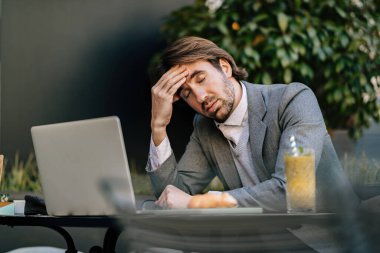 Exhausted entrepreneur holding his head in pain while sitting with eyes closed in outdoor cafe.