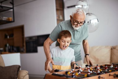 Happy little boy playing table football with his grandfather at home.