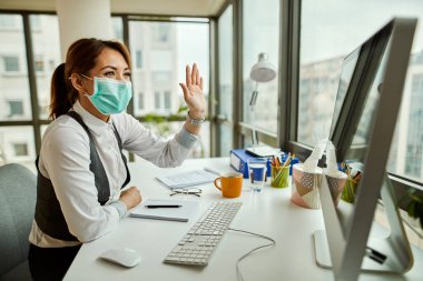Happy businesswoman with face mask using computer and waving to someone while making video call from her office. 