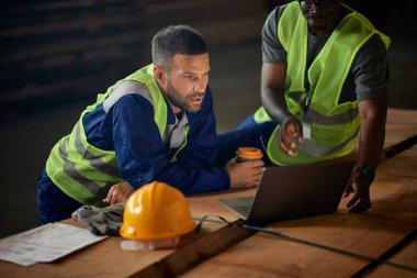 Warehouse workers reading delivery plans on laptop while working at lumber storage compartment. 