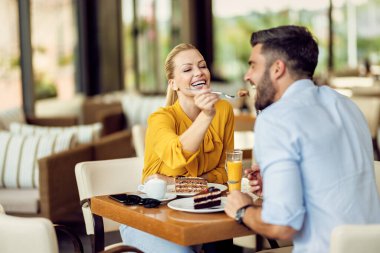 Happy woman having fun while feeding her boyfriend with a cake in a cafe.