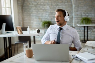 Mid adult businessman signing while listening music over headphones and working on laptop in the office. 