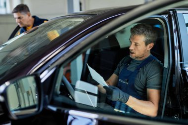 Auto mechanic using laptop and going through paperwork while sitting inside of a car in auto repair shop. 