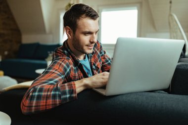 Smiling man surfing the net on laptop while relaxing on the sofa at home. 