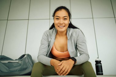 Low angle view of female Asian athlete relaxing in dressing room after sports training at the gym and looking at camera. 