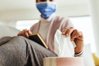 Close-up of woman with protective face mask taking a tissue from the box while reading book at home/