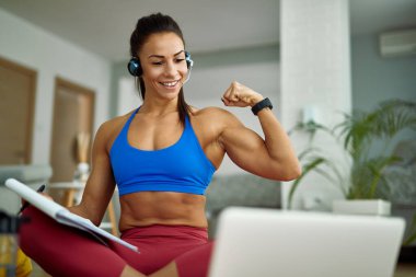 Young happy sportswoman flexing her bicep while making video call over laptop at home. 