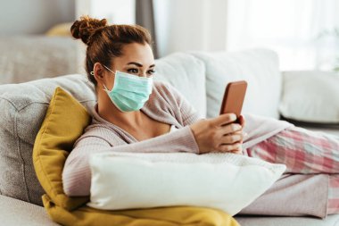 Smiling woman wearing protective face mask while using smart phone and text messaging at home. 