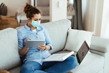 Freelance worker with face mask using digital tablet while going through business reports at home. 