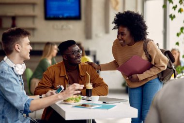 Happy university students communicating while enjoying on lunch break in cafeteria. Focus is on male black student. 