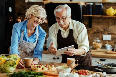 Happy senior couple surfing the net on touchpad while preparing food in the kitchen. 