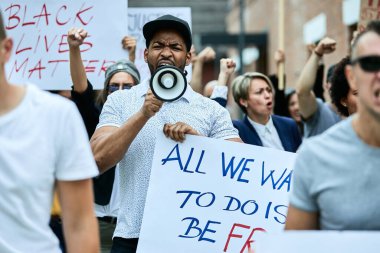 Multi-ethnic crowd of people protesting against racism on city streets. Focus is on African American man shouting through megaphone.