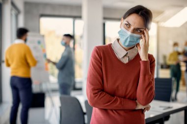Worried female entrepreneur with protective face mask thinking of something in the office. 