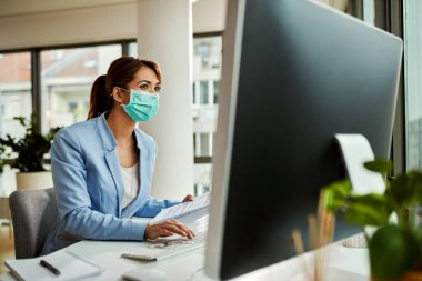Female entrepreneur wearing face mask while analyzing business reports and working on desktop PC in the office. 