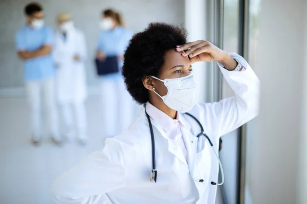 Distraught black doctor wearing protective face mask and thinking of something while looking through the window at medical clinic.