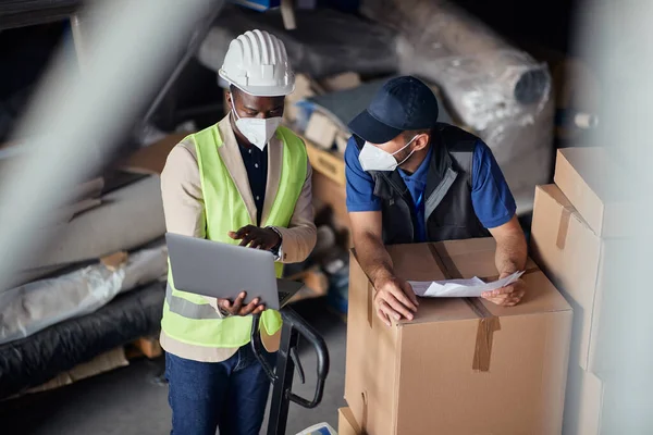 Black foreman and warehouse worker talking while using laptop and wearing protective face masks storage compartment.