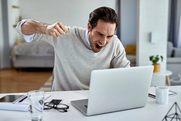 Angry businessman shouting from frustration and about to punch his laptop after reading bad news while working at home.