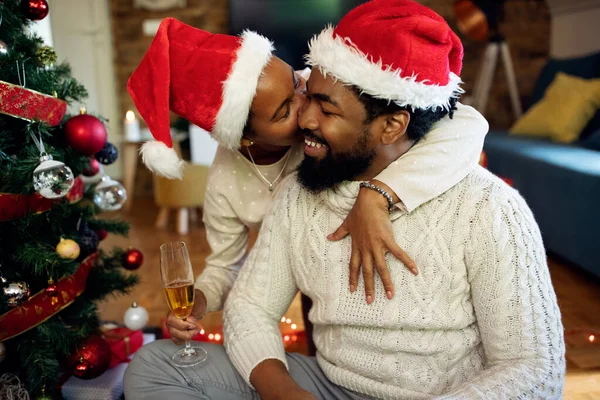 Happy African American man being kissed by his wife while celebrating Christmas together at home.