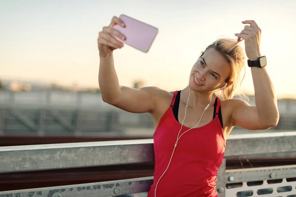 Smiling athletic woman using mobile phone and taking selfie while working out outdoors.