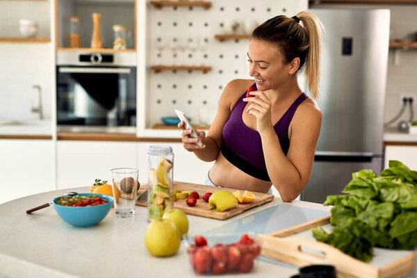Young happy athletic woman eating fruit while text messaging on mobile phone in the kitchen. 