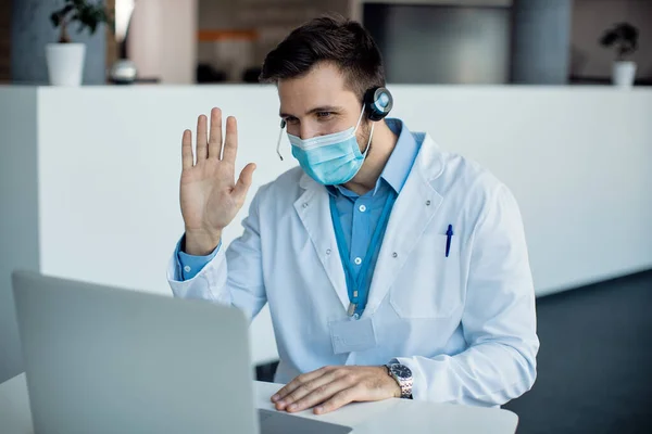 Doctor with face mask greeting someone while having video call over laptop in the hospital.