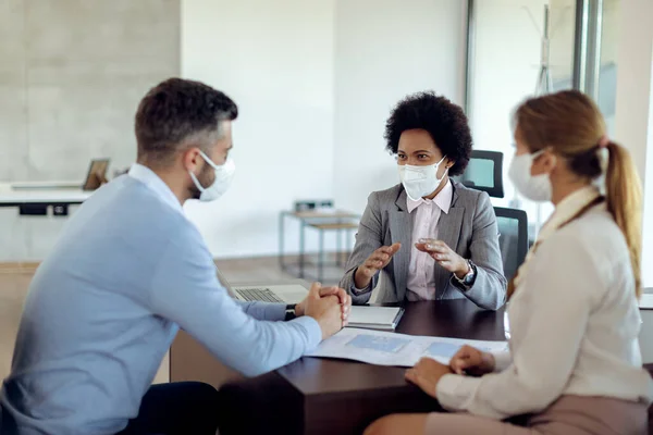 Black real estate agent and a couple wearing protective face masks while communicating during the meeting in the office.