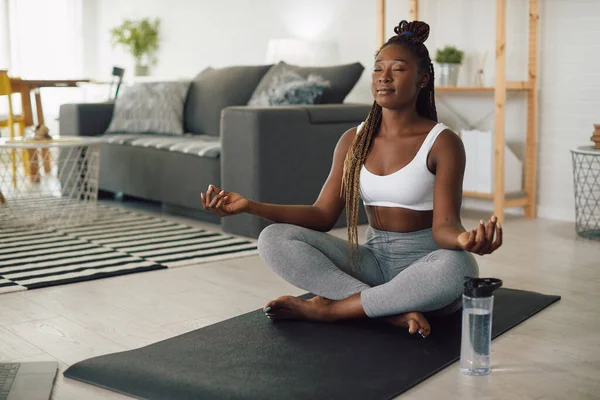 Black athletic woman doing breathing exercises in lotus position while practicing Yoga in the living room.