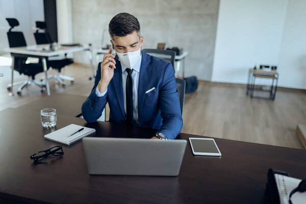 Businessman wearing protective face mask while working on laptop and communicating over cell phone in the office.
