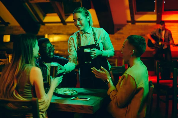 Young happy waitress serving beer to group of young people who are having fun at night in a bar.