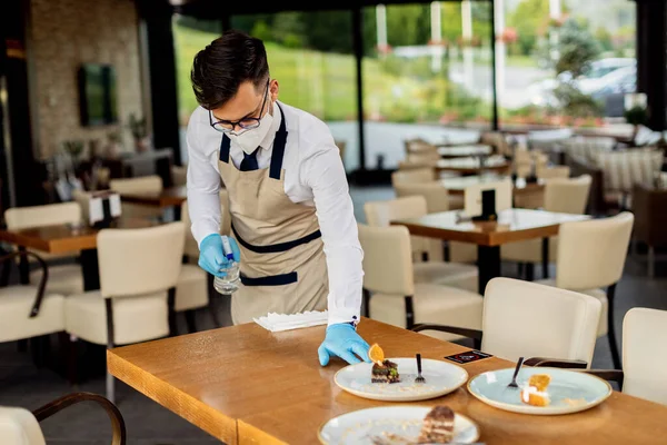 Waiter with face mask disinfecting table after the guests in a cafe during coronavirus epidemic.