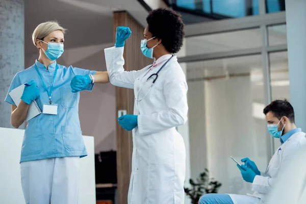 Happy nurse and female doctor greeting with elbows in a hallway at hospital during COVID-19 pandemic.