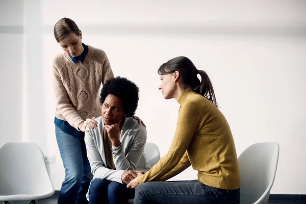 Sad African American woman being consoled by female participants of group therapy at community center.