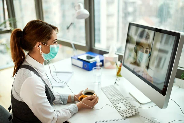 Smiling businesswoman with face mask using computer and making video call while having coffee break in the office.