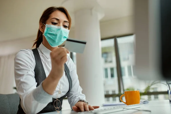 Businesswoman with protective face mask using computer and credit card while e-banking in the office.