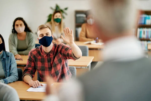 Male student raising hand to answer teacher\'s question and wearing protective face mask due to coronavirus pandemic.