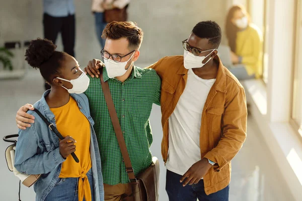 Small group of happy college friends with protective face masks embracing while standing in a lobby and talking.