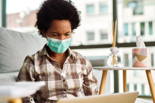 Black woman with face mask using computer while working at home during virus epidemic.