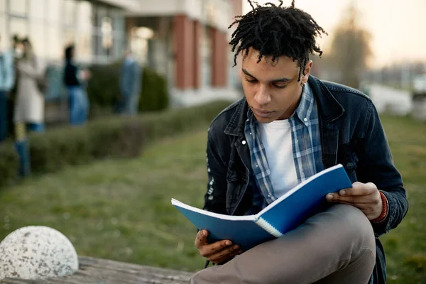 Black student learning from his textbook while sitting outdoors at campus.