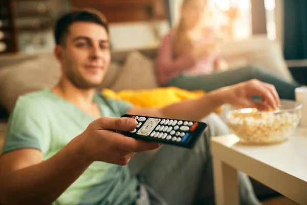 Close-up of man using remote controller and changing channels while watching TV at home.