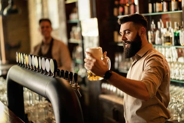 Young happy bartender holding a glass of draft beer while working at bar counter. 
