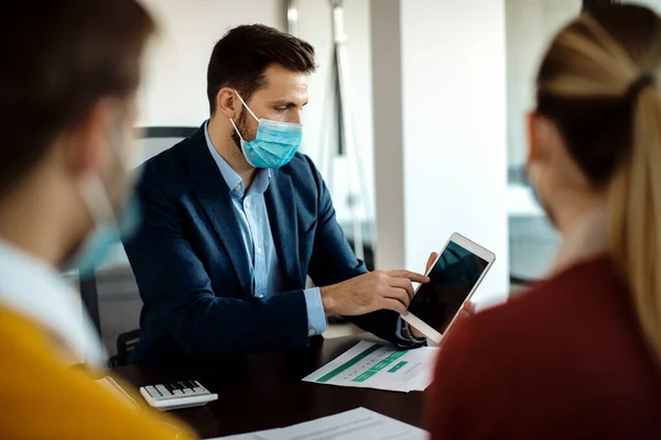 Financial consultant wearing protective face mask while having a meeting with clients and working on digital tablet in the office.