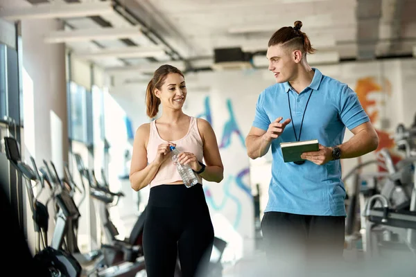 Happy athletic woman and her personal trainer talking while walking through gym during sport training.