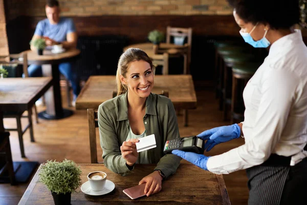 Happy woman using credit card while making contactless payment to a waitress due to coronavirus pandemic.