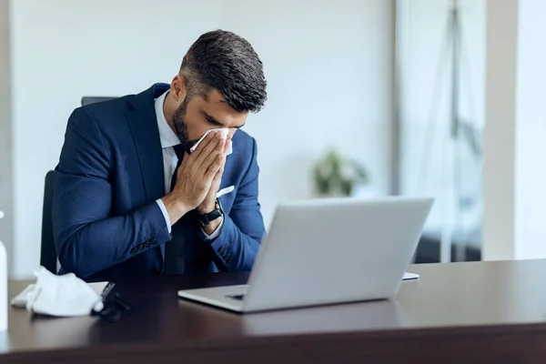 Young businessman feeling ill and sneezing in a tissue while working on laptop in the office.