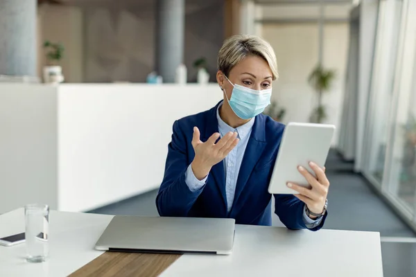 Female entrepreneur using digital tablet while having conference call and wearing protective face mask due to coronavirus pandemic.
