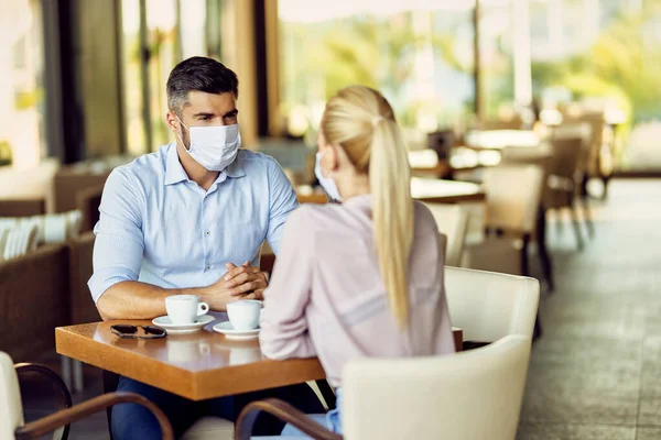 Couple with face masks relaxing in a cafe and communicating. Focus is on man.